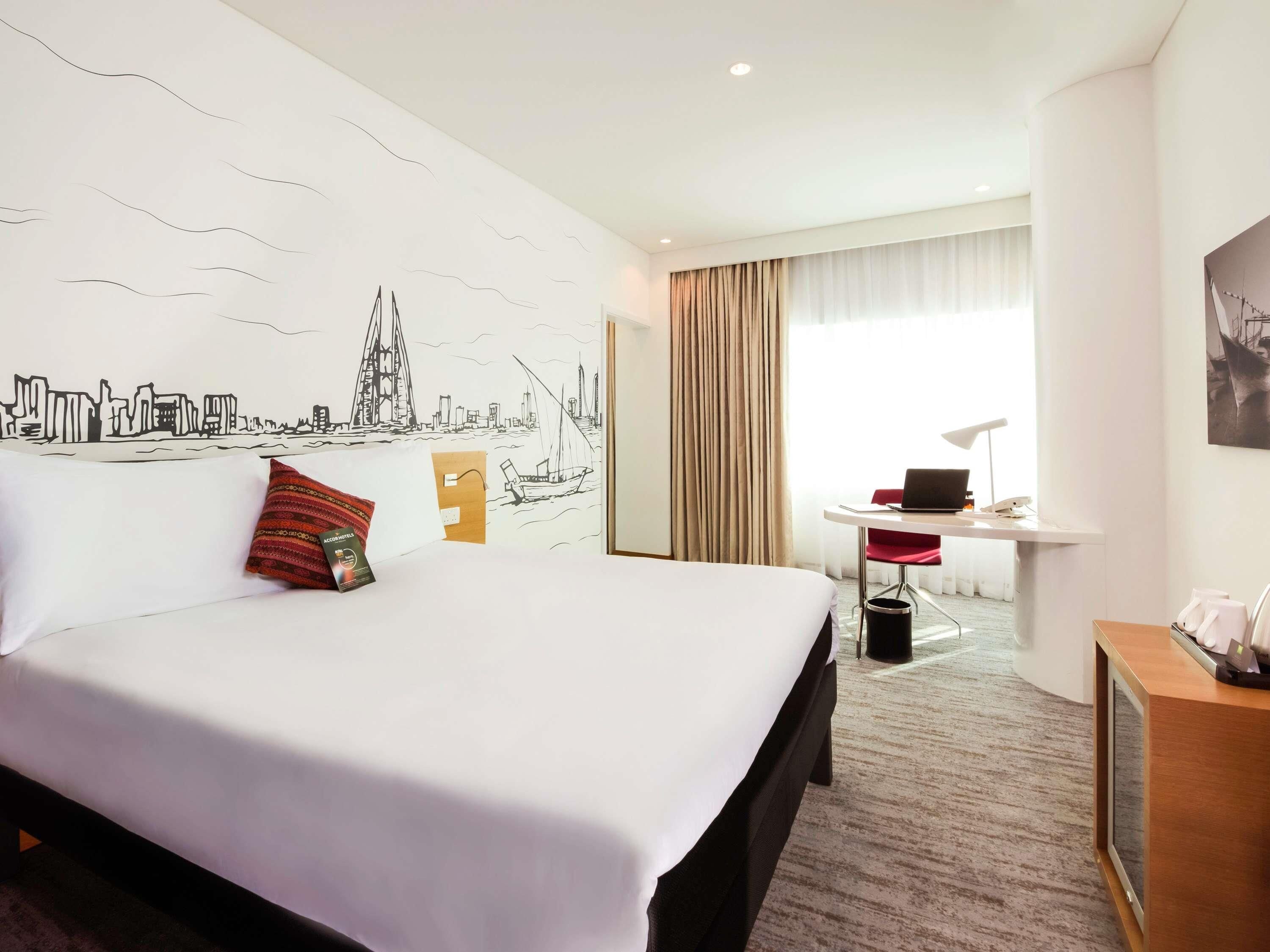 A hotel room with a large bed, a desk with a laptop, a chair, and a window with curtains.