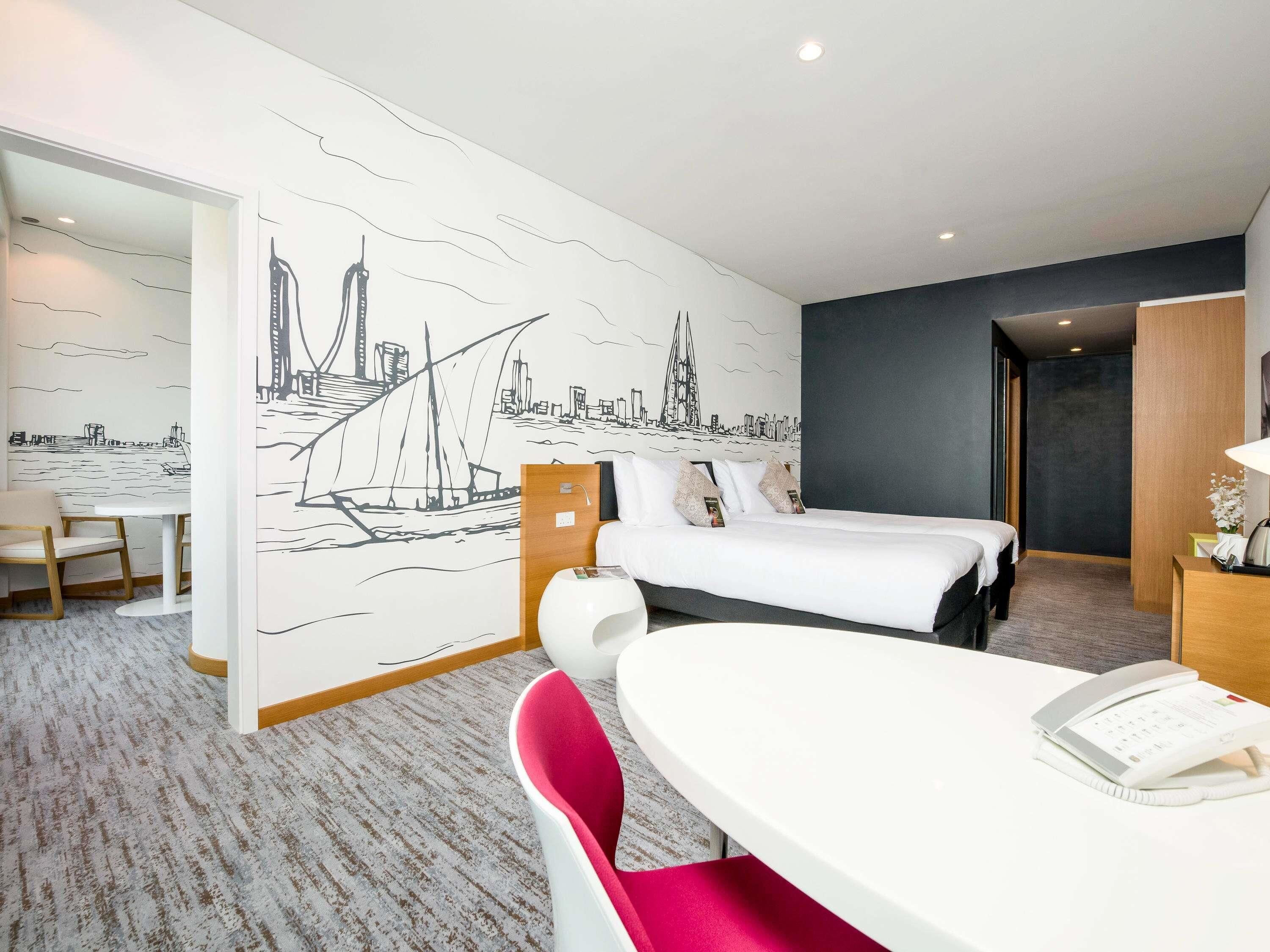Habitación de hotel con cama, escritorio, silla y un mural en la pared que representa un paisaje urbano.