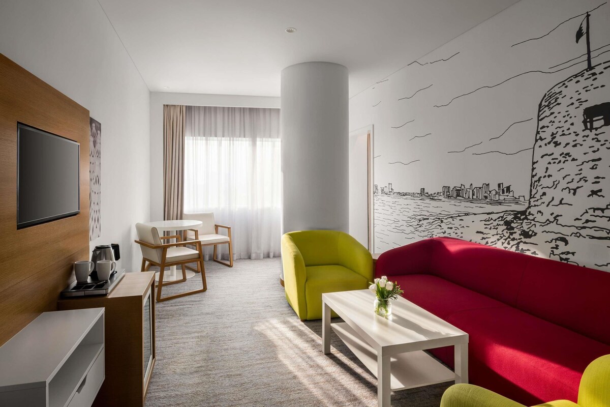 A modern hotel room with a flat-screen TV, a dining area with a table and chairs, a vibrant red sofa, a yellow armchair, and a mural of a city skyline on the wall.