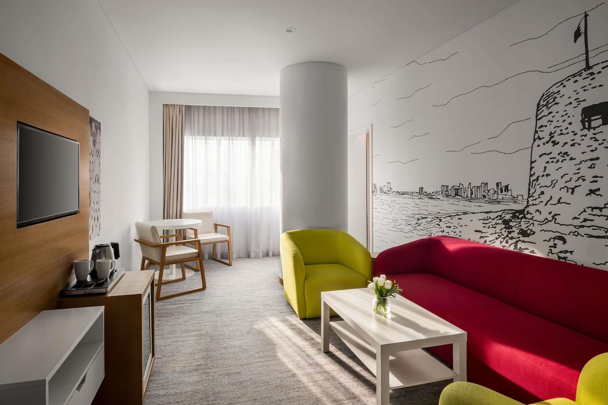 A modern hotel room with a flat-screen TV, a dining area with a table and chairs, a vibrant red sofa, a yellow armchair, and a mural of a city skyline on the wall.