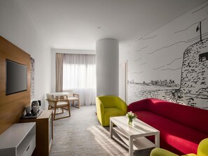 Una habitación de hotel moderna con un televisor de pantalla plana, una zona de comedor con mesa y sillas, un sofá rojo vibrante, un sillón amarillo y un mural con un paisaje urbano en la pared.