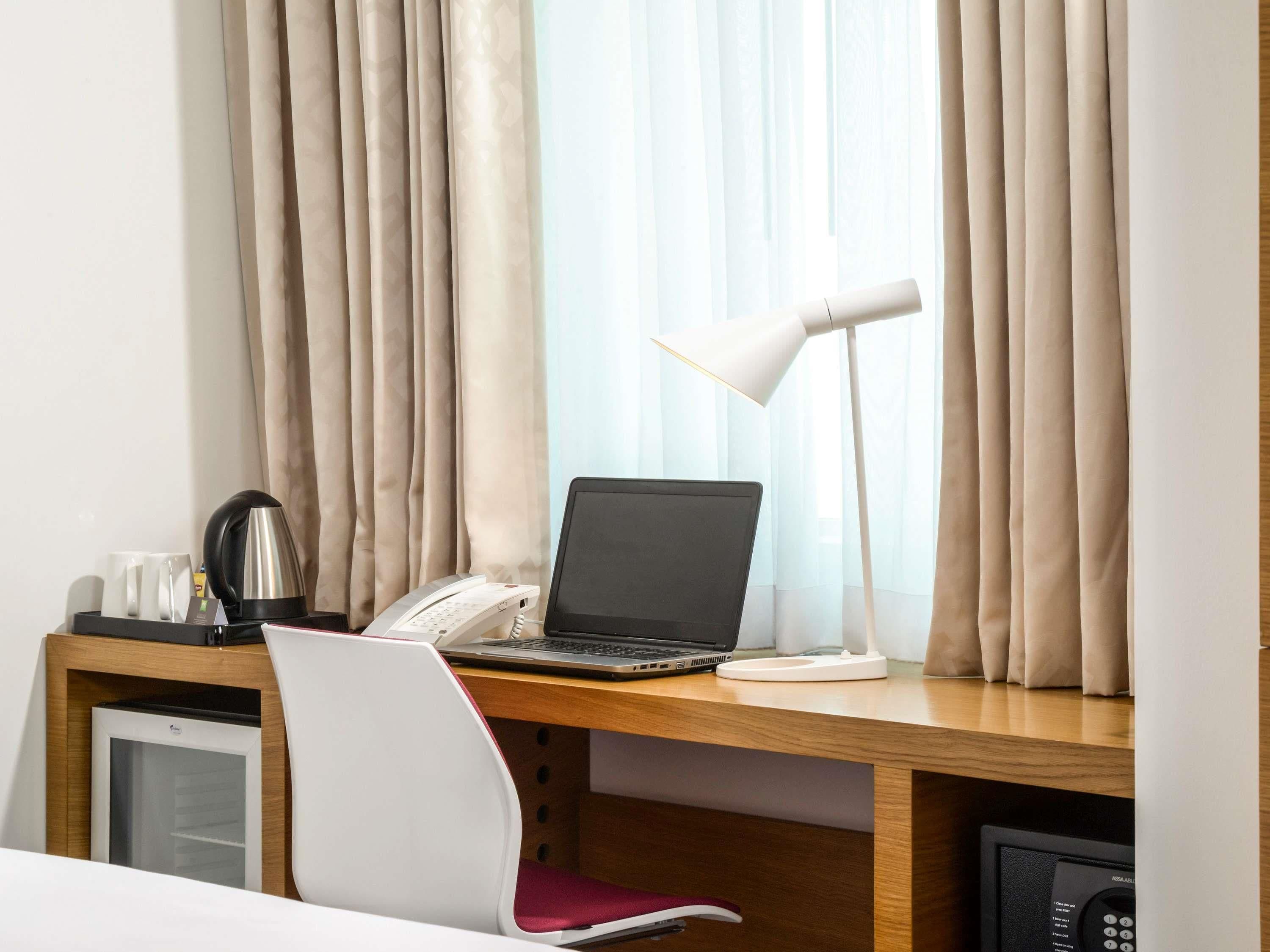 A hotel room with a wooden desk, a laptop, a telephone, a coffee maker, and a mini-fridge.