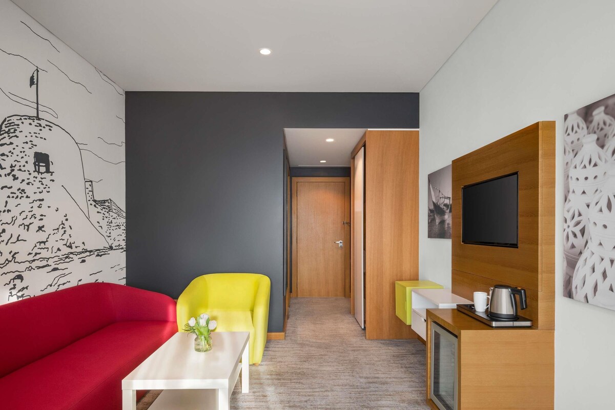 A modern hotel room with a red sofa, a yellow armchair, a small white table, a TV, and a wooden cabinet with a kettle.