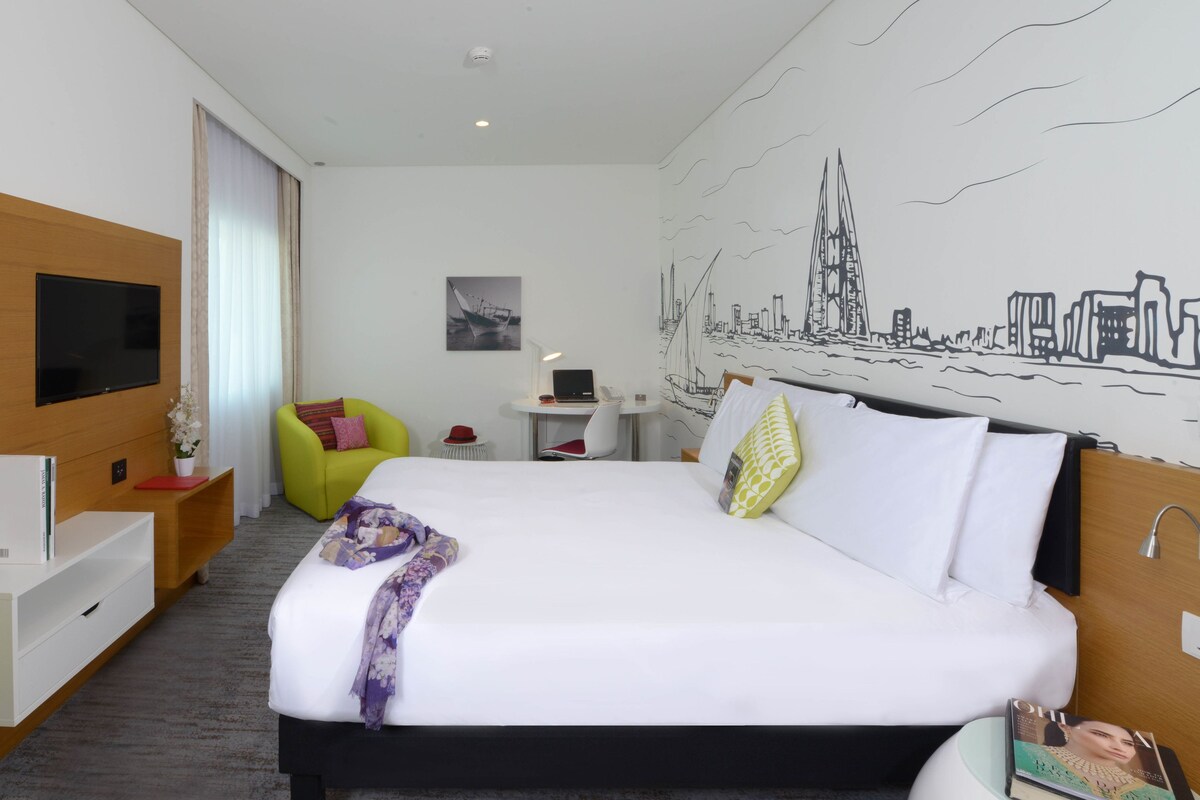 Habitación de hotel con cama, escritorio, silla, televisor y un mural en la pared.