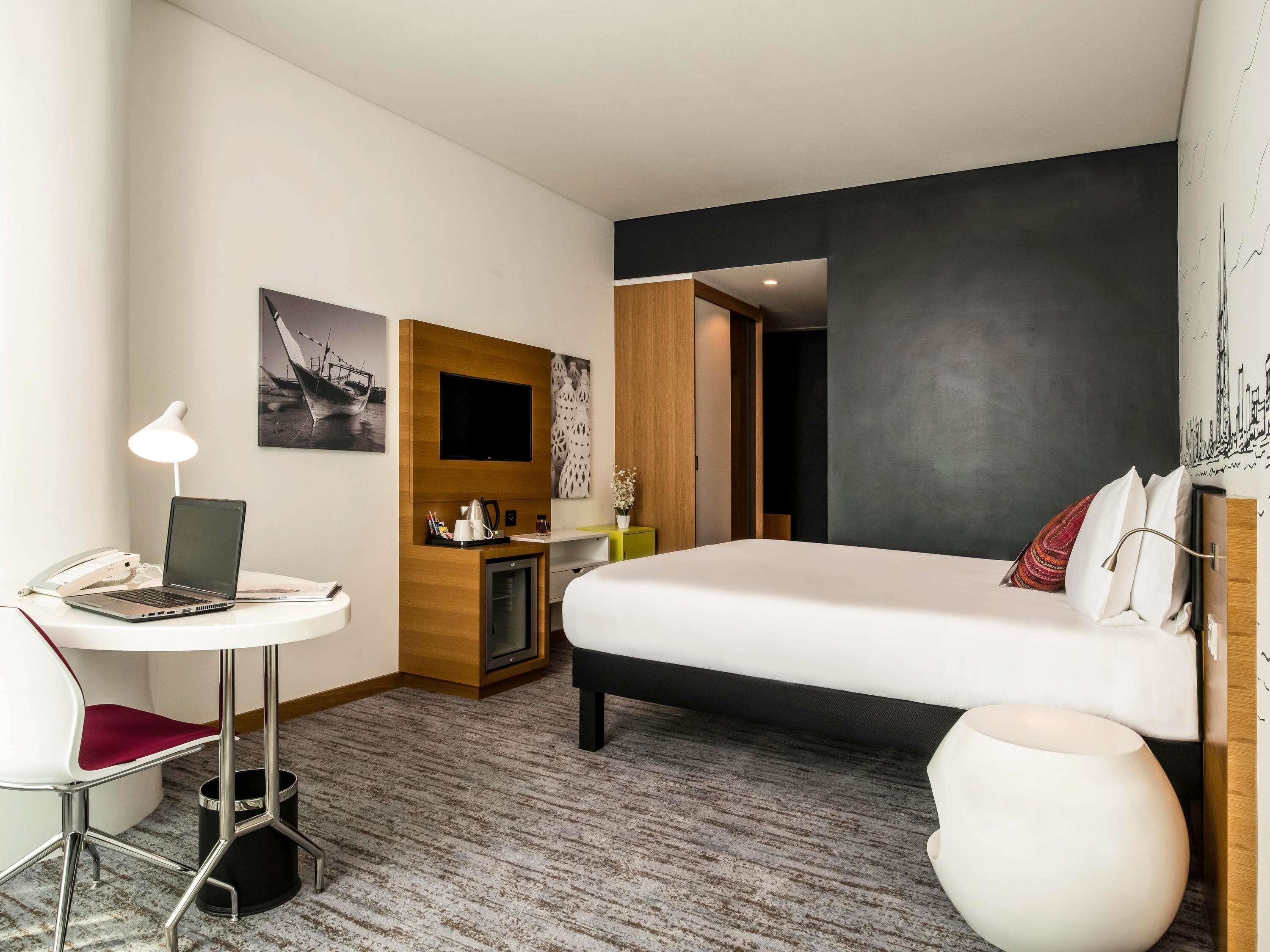 Ein Hotelzimmer mit einem Bett, einem Schreibtisch mit Laptop, einem Sessel, einem kleinen Tisch, einem Papierkorb und einem an der Wand befestigten Fernseher.