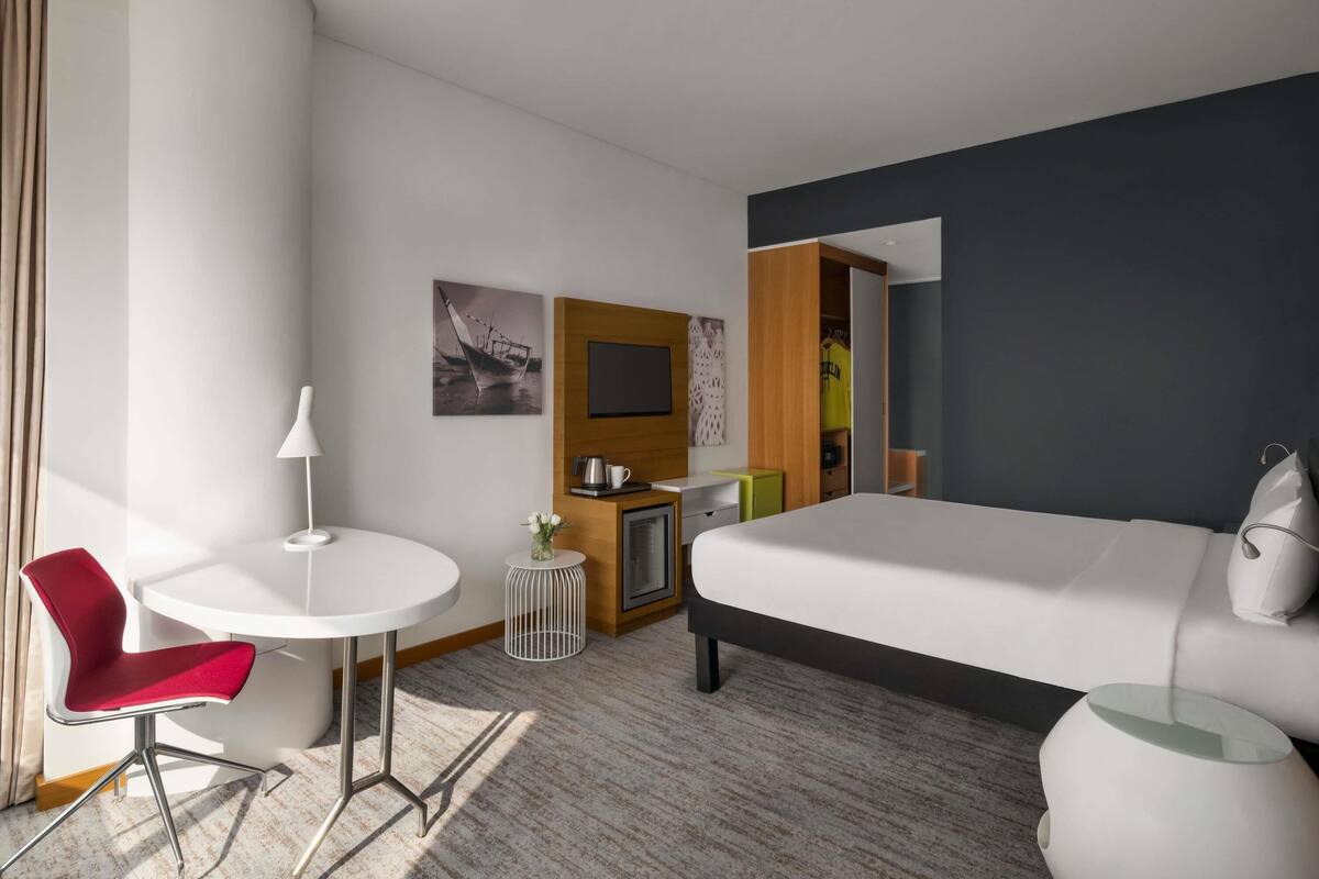 A modern hotel room with a bed, a desk with a chair, a small round table, and a mirror.