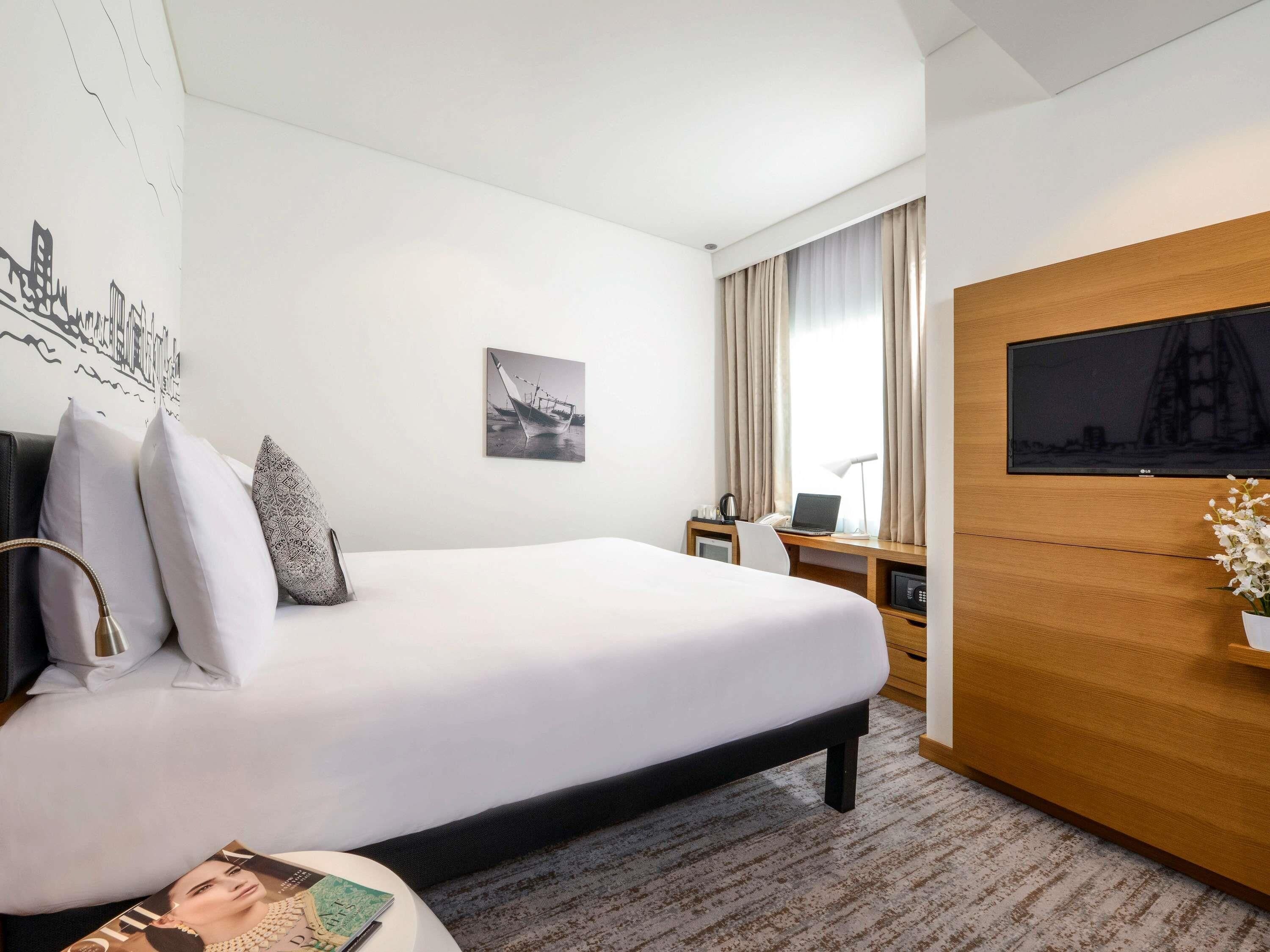 A hotel room with a bed, a desk with a laptop, a TV, and a picture of a boat on the wall.