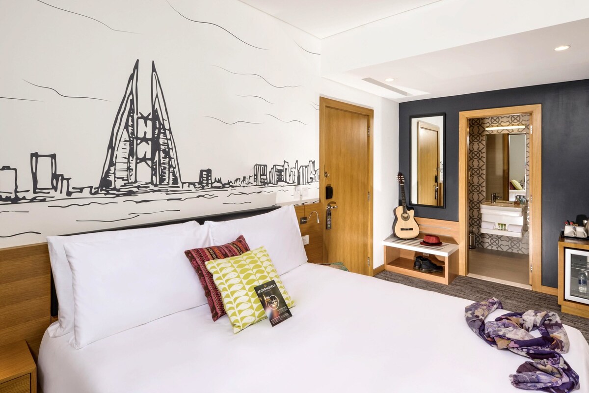 Habitación de hotel con una cama, una guitarra, un tocador con espejo y un mural urbano en la pared.