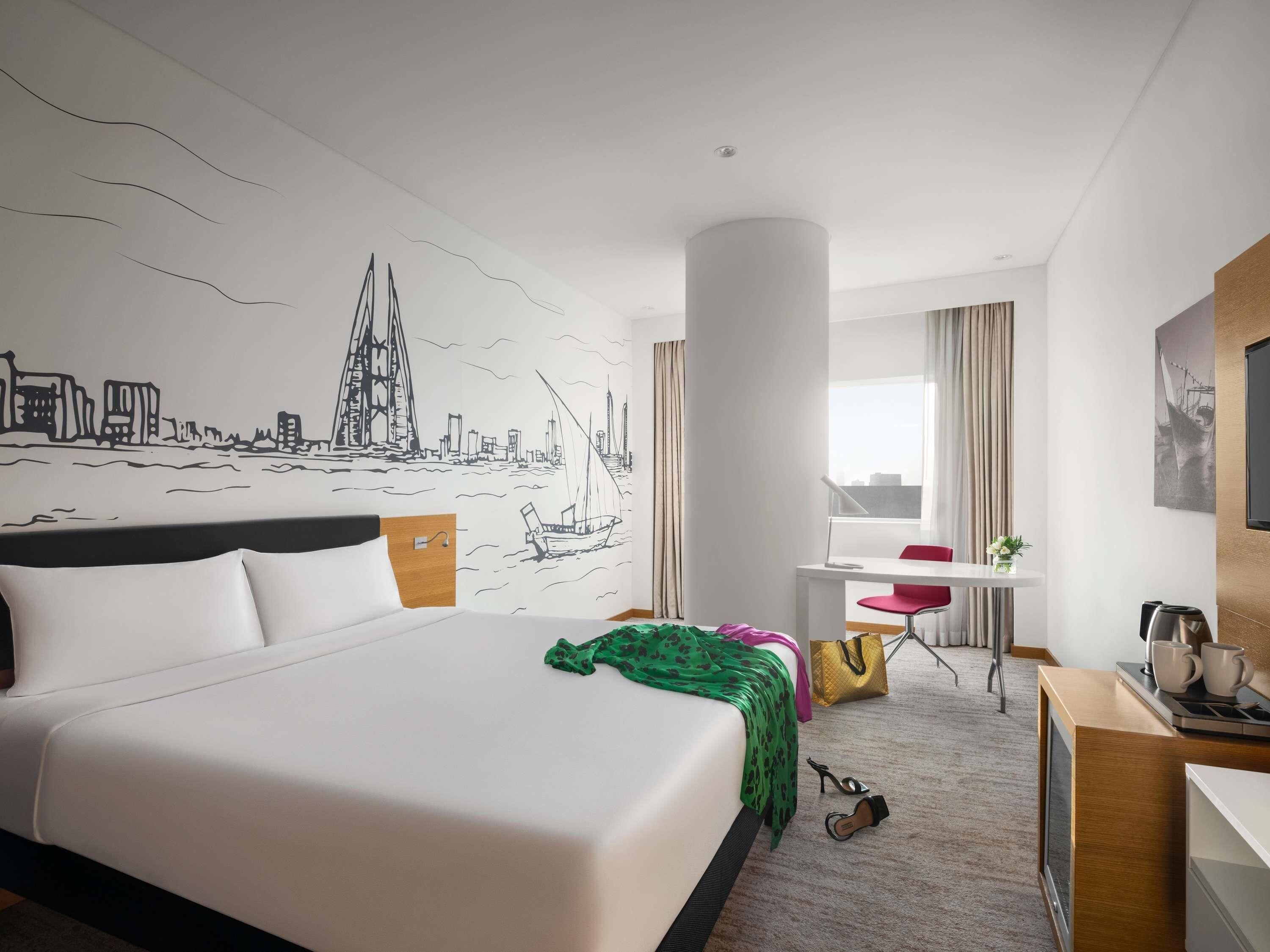 Habitación de hotel con una cama grande, un escritorio con silla, un televisor y un mural con vistas a la ciudad.
