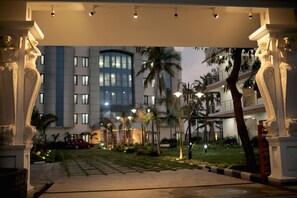 A hotel entrance with classical columns, a well-lit pathway, palm trees, and a modern building in the background. - Lee Crysstal (Tirupporur)