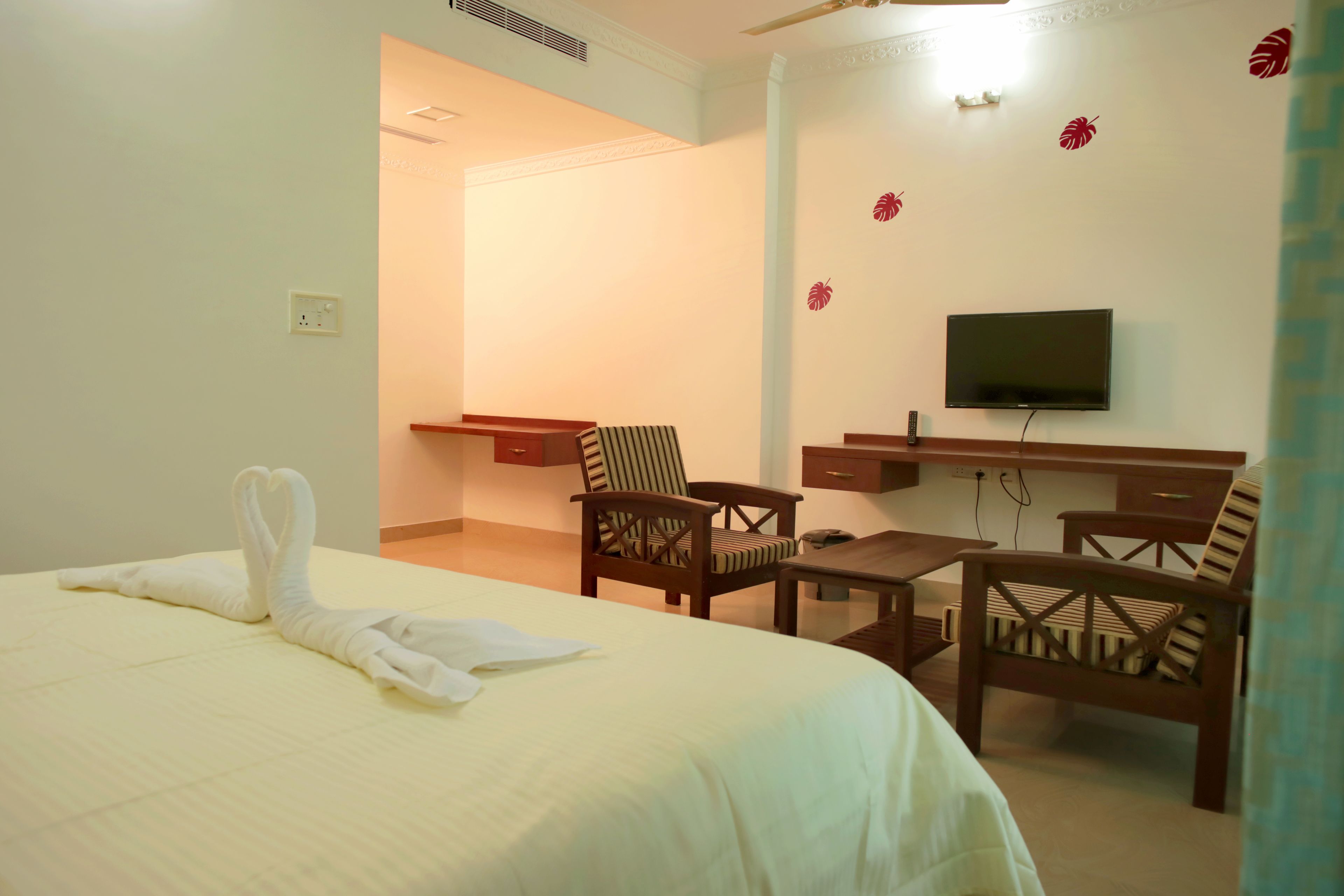 A hotel room with a bed, two wooden chairs, a small table, a desk, a television, and a wall with leaf decals.