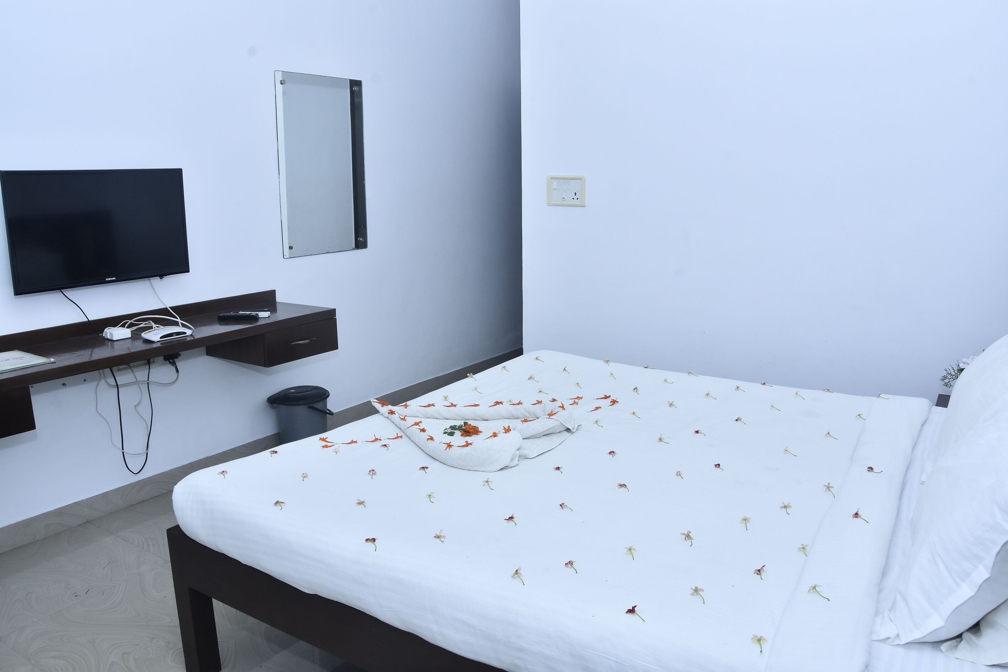 Una cama bien hecha con una colcha estampada con motivos florales, un televisor montado en la pared y un cubo de basura pequeño al lado de la cama.
