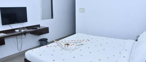 Una cama bien hecha con una colcha estampada con motivos florales, un televisor montado en la pared y un cubo de basura pequeño al lado de la cama.