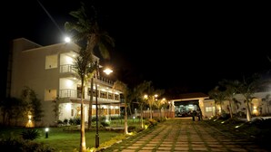 A well-lit hotel entrance with a paved pathway, palm trees, and a modern building facade. - Lee Crysstal (Tirupporur)