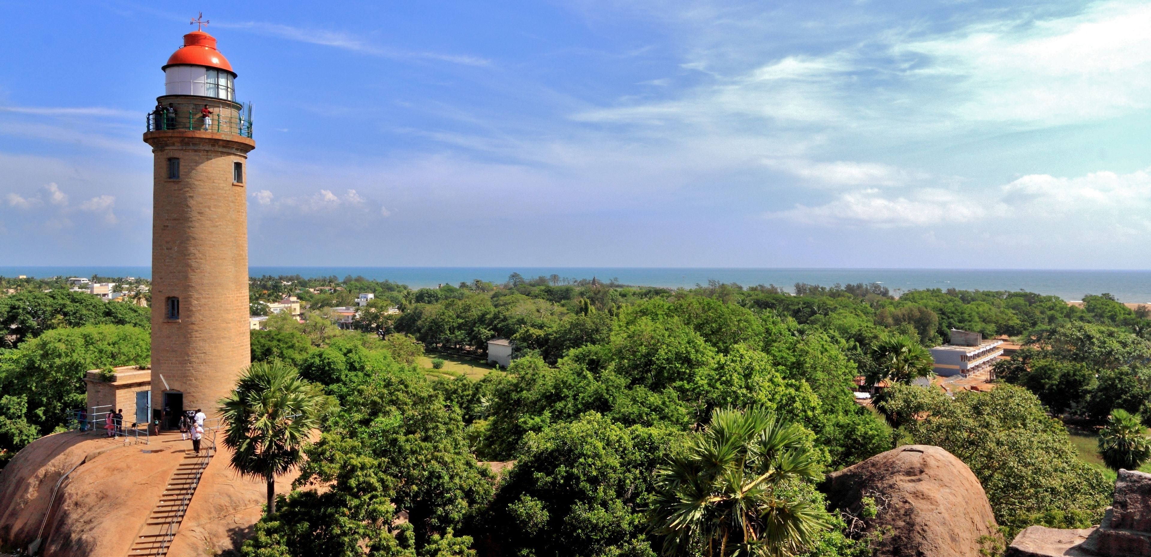 A lighthouse with a brick base and a red top, surrounded by lush greenery and a clear sky.