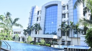 A modern hotel with a swimming pool, palm trees, and a large blue glass facade. - Lee Crysstal (Tirupporur)