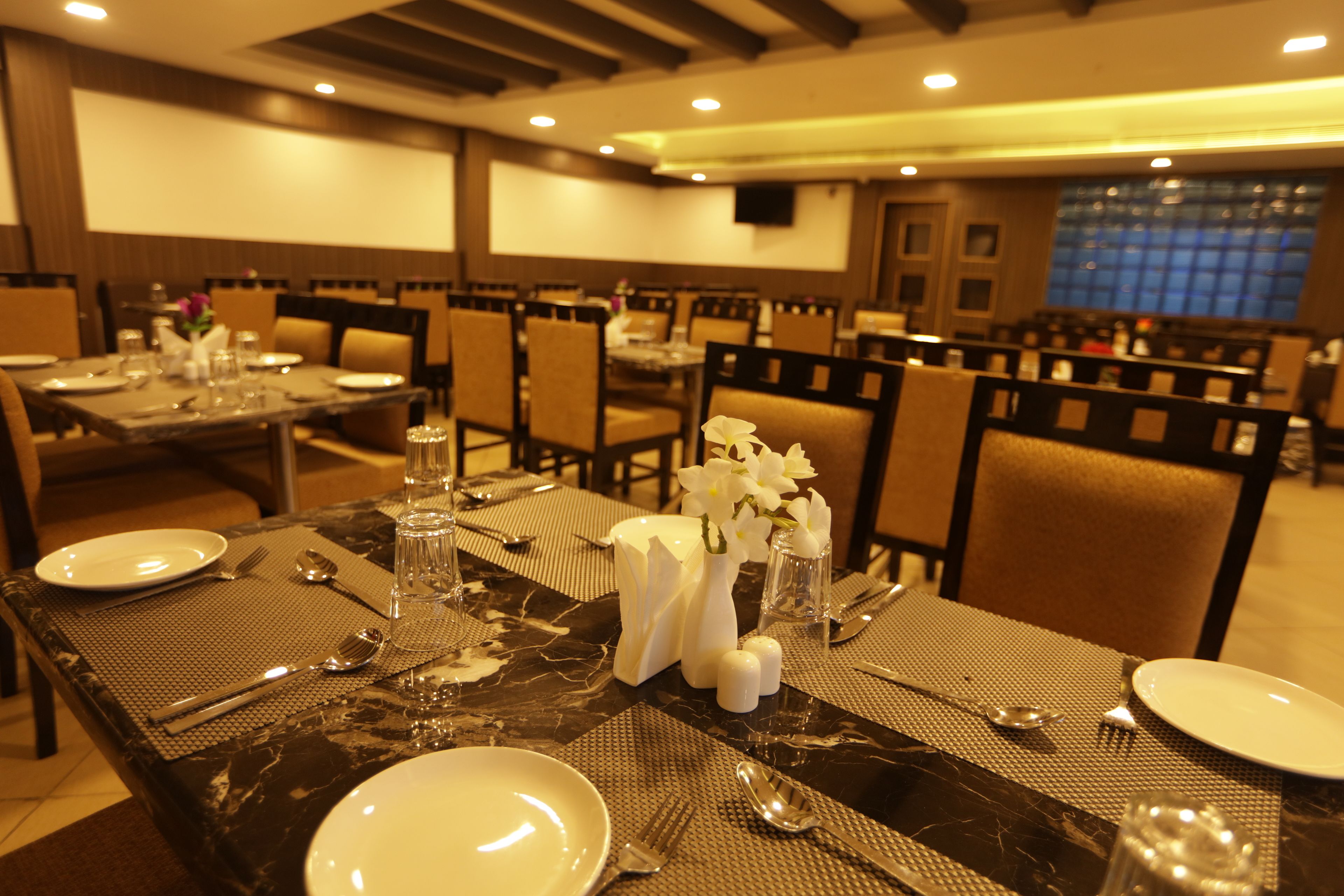 A dining area with tables set for four, each with a marble tablecloth, white plates, cutlery, and a vase with white flowers.