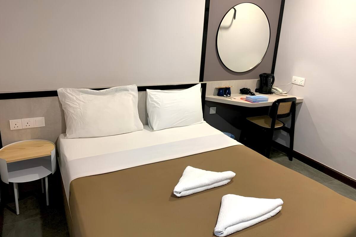 A hotel room with a bed, two pillows, a desk with a chair, a round mirror, and a small table with a chair.