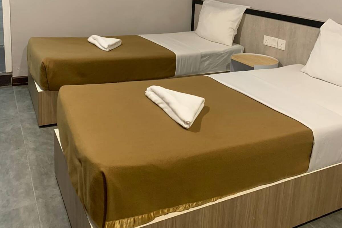 A hotel room with two beds, each with a brown bedspread and white pillows, a small bedside table with a lamp, and an air conditioning unit on the wall.