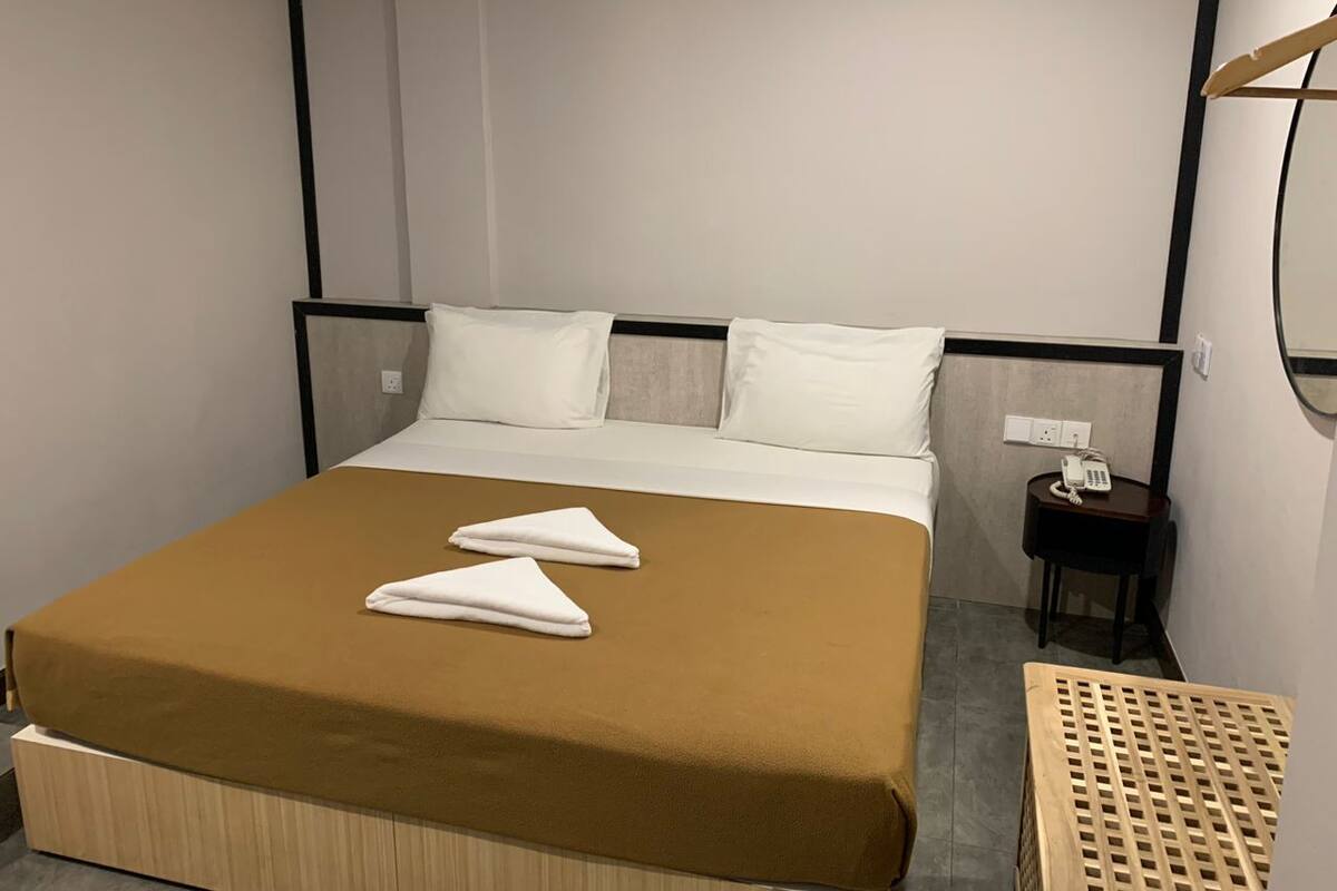 A hotel room with a bed, two white towels, a wooden nightstand with a phone, and a small table with a grid pattern.