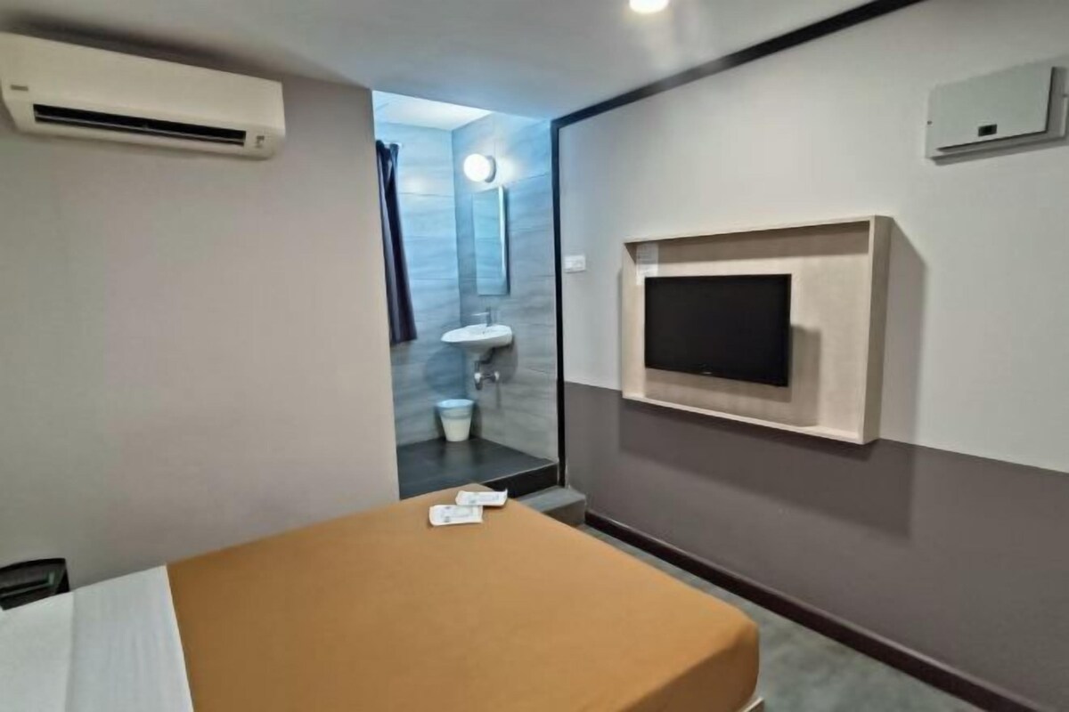 A hotel room with a bed, a TV mounted on the wall, an air conditioning unit, and a bathroom visible through an open door.