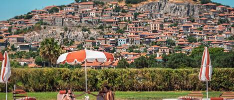 A pool area with lounge chairs and umbrellas, a clear view of a historic hilltop with buildings, and a large body of water.