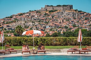 A pool area with lounge chairs and umbrellas, a clear view of a historic hilltop with buildings, and a large body of water. - Hotel Delfinia (Lesvos)