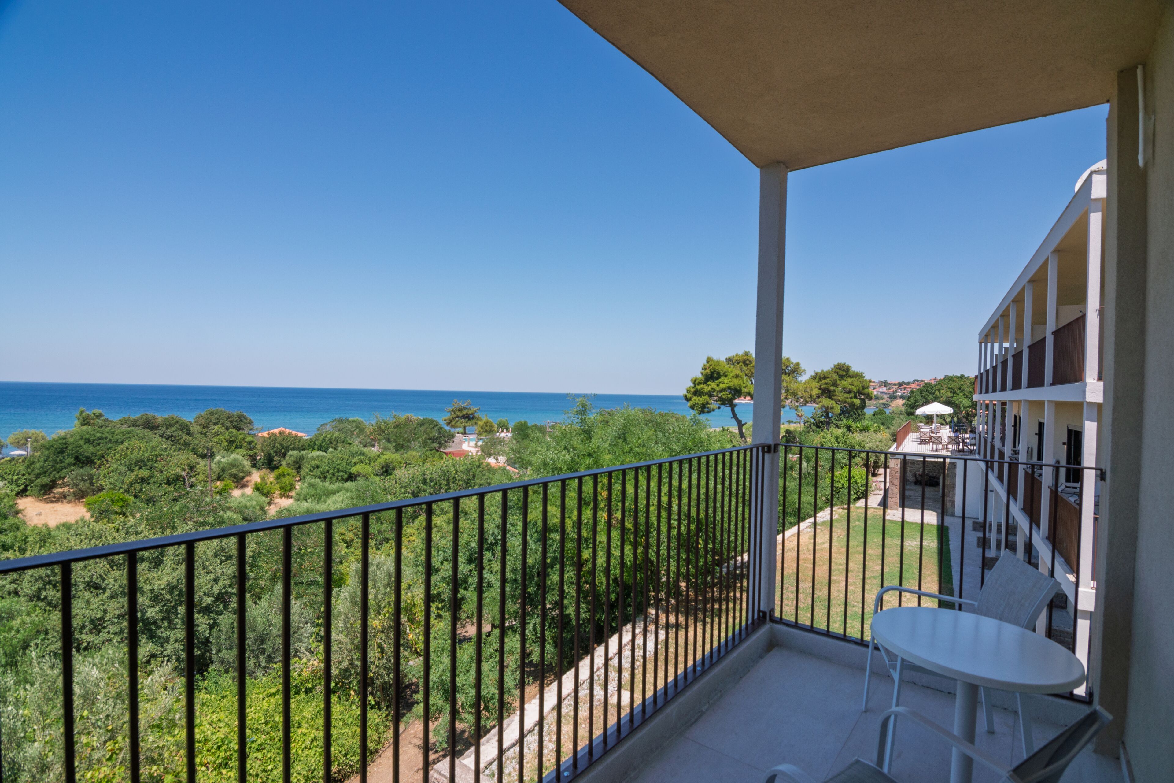 A balcony with a table and chairs, overlooking a coastal landscape with trees and a clear sky.
