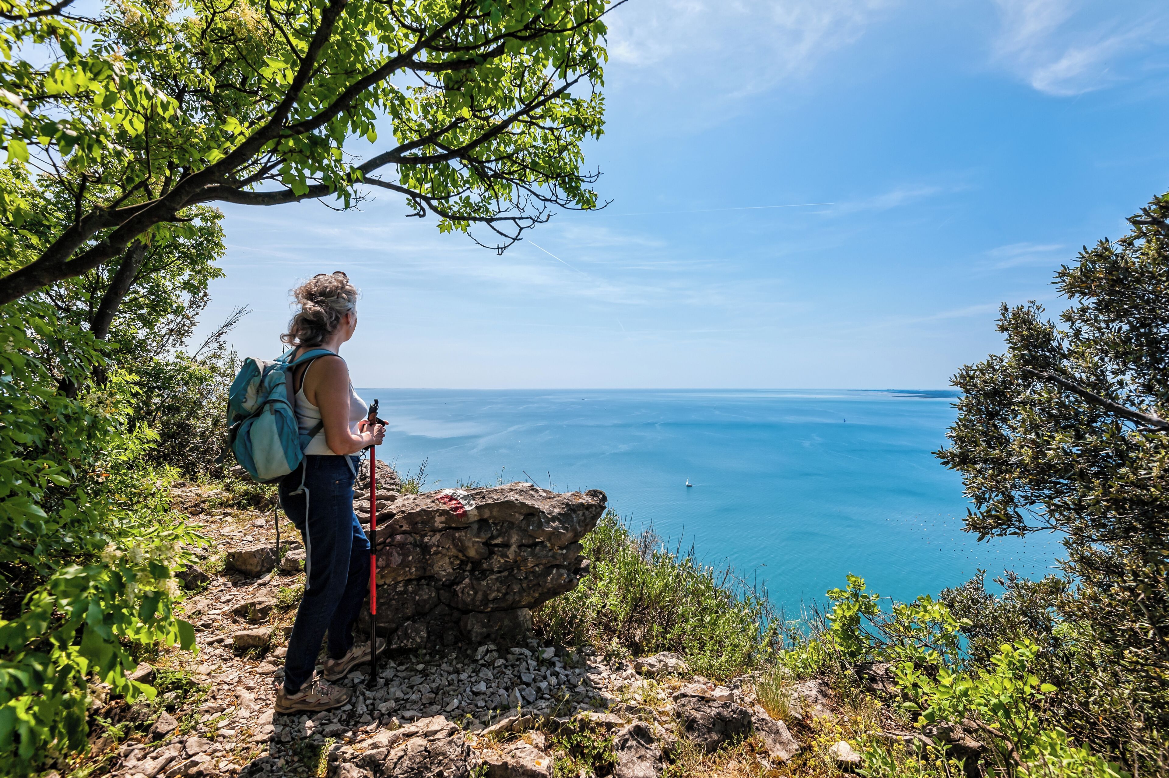 A hiker on a rocky path with a backpack, overlooking a clear blue sea.