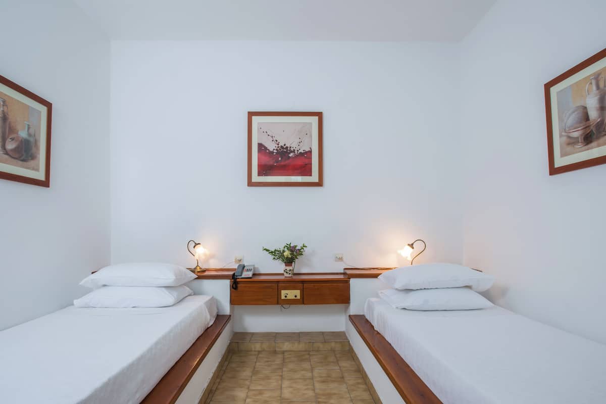 A room with two single beds, a wooden nightstand with a telephone, a vase with flowers, and framed artwork on the walls.