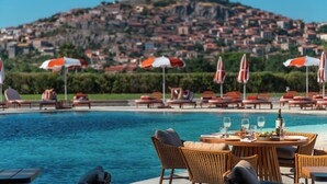 Un área junto a la piscina con sillones de descanso y sombrillas, una mesa de comedor para dos personas, y vista a una colina con edificios.
