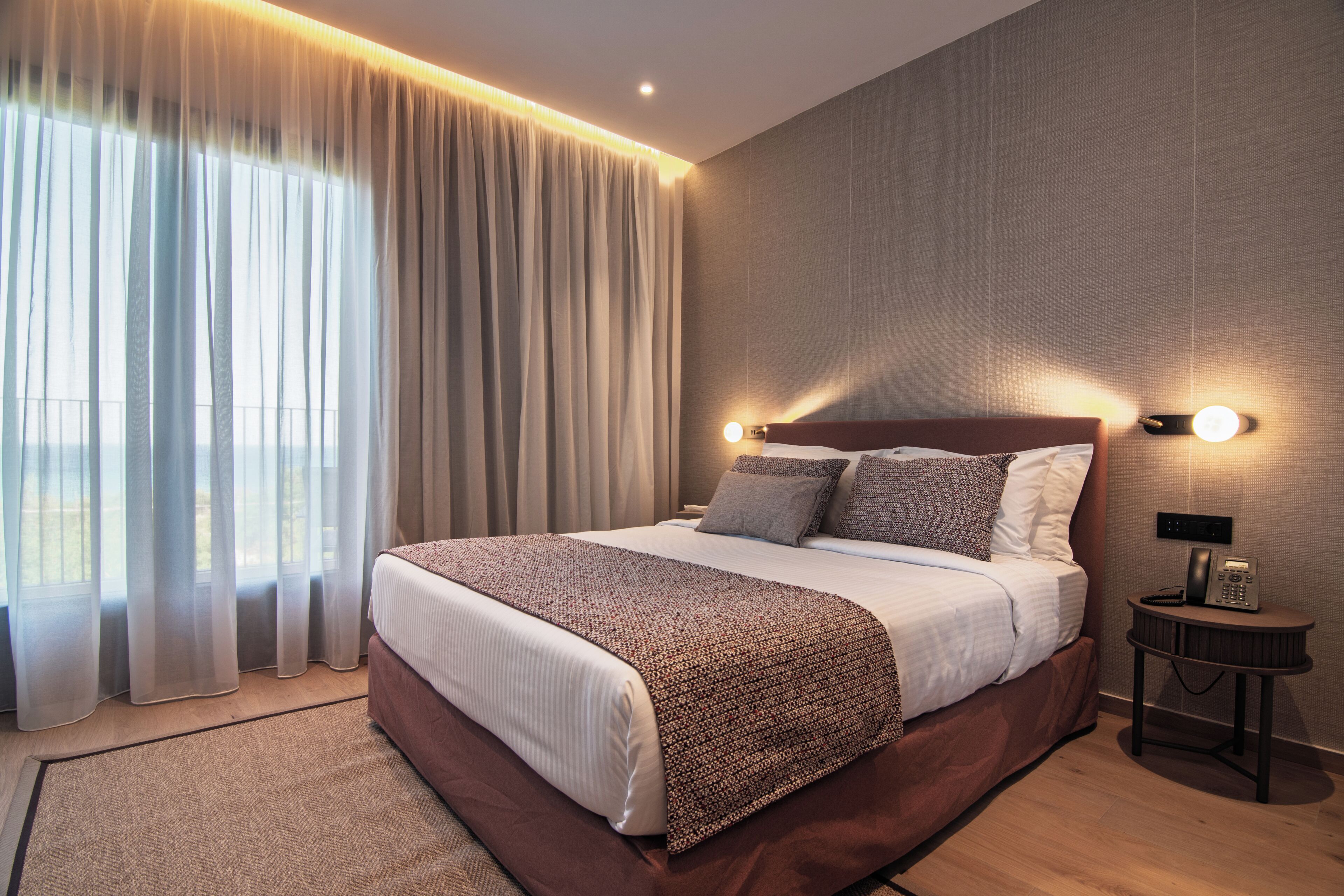 Une chambre d’hôtel moderne avec un grand lit, une table de chevet avec un téléphone et une vue sur l’extérieur à travers des rideaux légers.