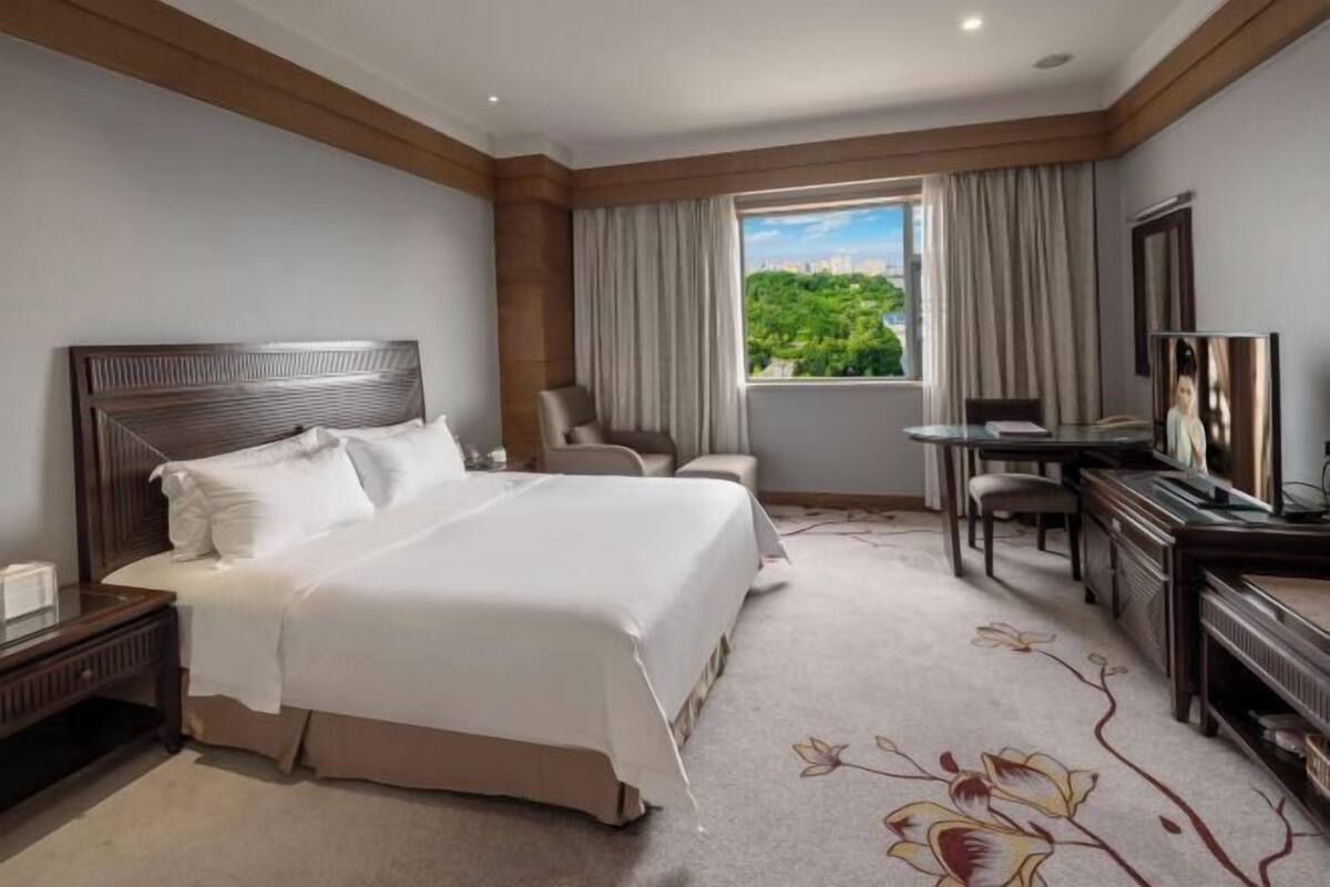 A hotel room with a large bed, a desk with a chair, a television, and a view of greenery outside.