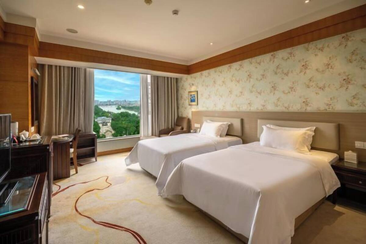 A hotel room with two beds, a desk with a computer, a chair, and a large window with a city view.