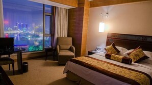 A hotel room with a large window offering a city view, a bed with a patterned bedspread, a chair, a desk, and a nightstand with a lamp. - Eyring Daqian International Hotel (Neijiang City)