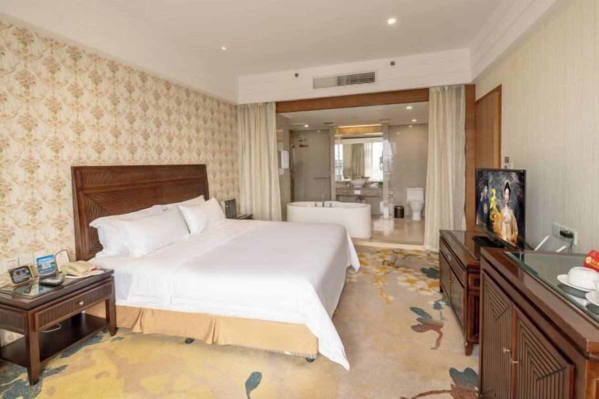 A hotel room with a large bed, a bedside table with a phone, a mirror, a bathroom with a bathtub, and a TV on a wooden cabinet.