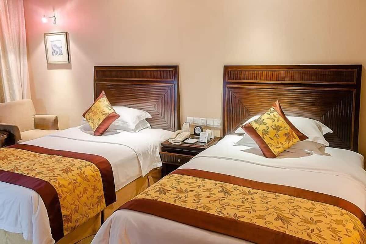 A hotel room with two beds, each with a wooden headboard, white bedding, and a floral-patterned bedspread. There is a nightstand with a telephone and a clock between the beds.