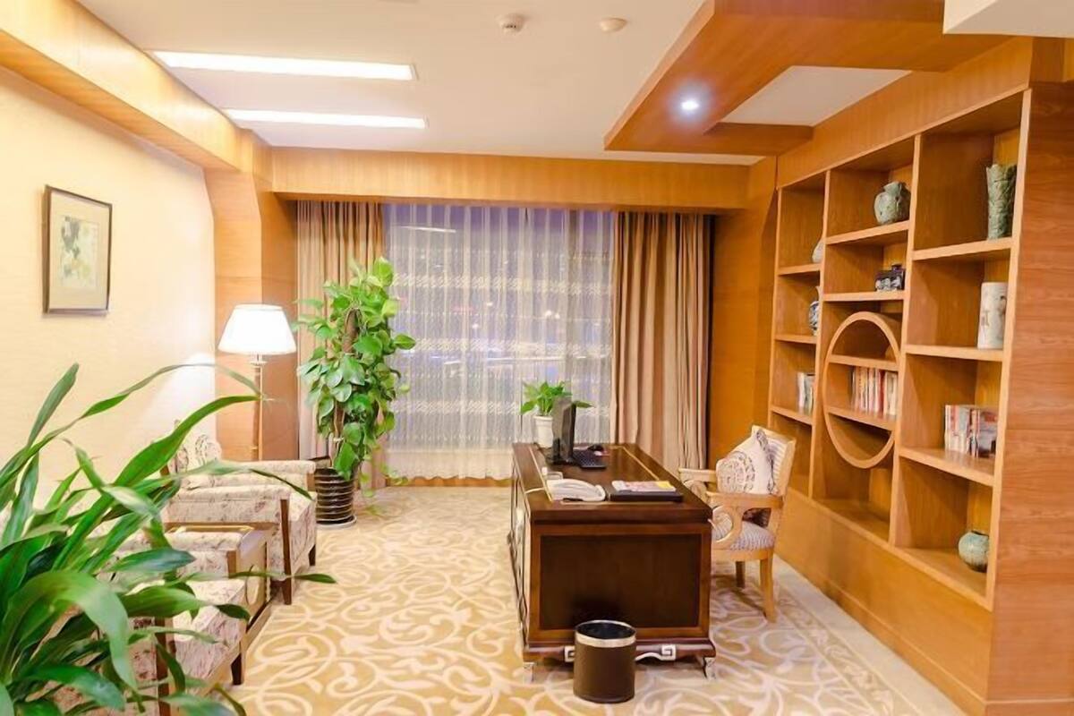 A hotel room with a desk, chair, bookshelf, large window with curtains, and a patterned carpet.