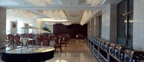 A spacious hotel lobby with a long row of tables and chairs, a central round table with a glass display, and a modern chandelier.