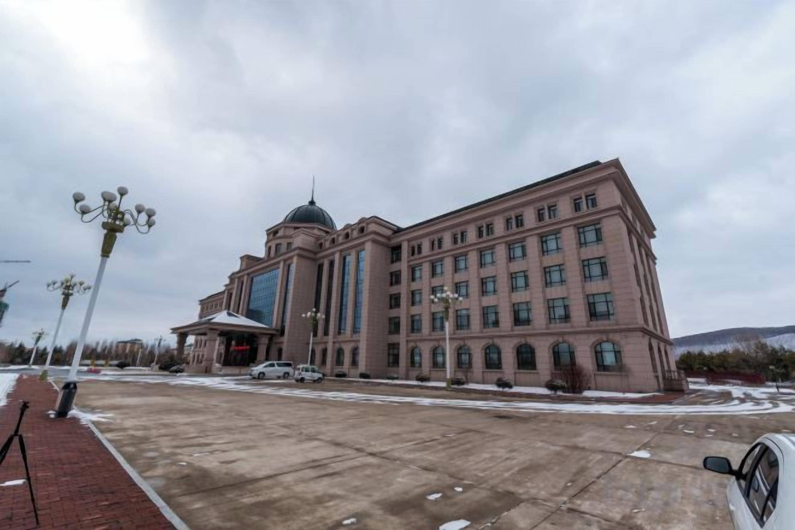 Un grand bâtiment doté d’un dôme central et de nombreuses fenêtres, une zone pavée devant et quelques véhicules.