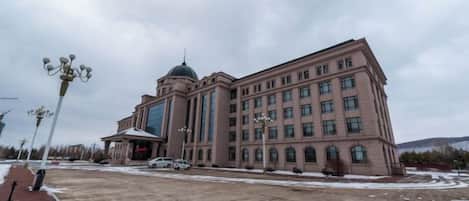 A large building with a central dome and multiple windows, a paved area in front, and a few vehicles.