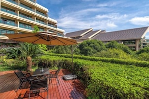 Un patio exterior con una mesa y sillas, rodeado de abundante vegetación y un gran paraguas.