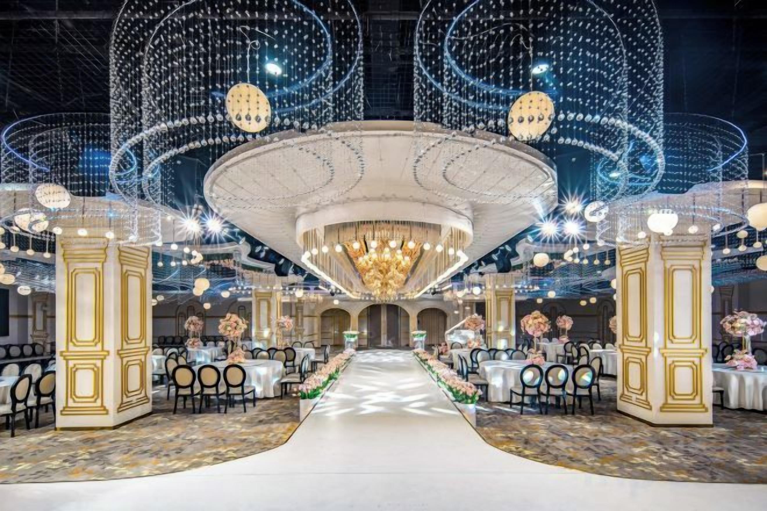 Un salón amplio con un pasillo central, columnas ornamentadas y candelabros. Mesas dispuestas a ambos lados del pasillo.