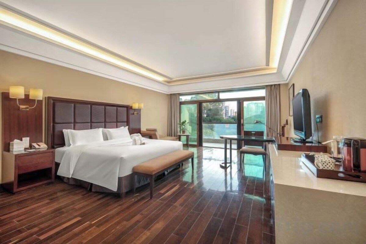 A hotel room with a large bed, bedside tables, a bench, a TV, and a balcony with a view.