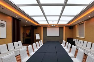 A conference room with a long table, white chairs, a projector screen, and a ceiling with a skylight.