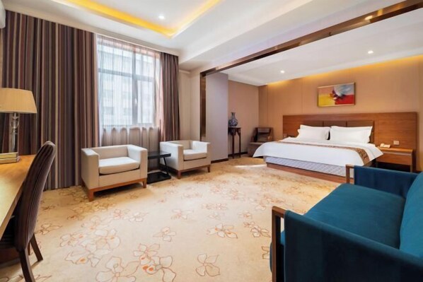 Une chambre d’hôtel avec un lit, deux fauteuils, un bureau avec une lampe et une grande fenêtre avec des rideaux.