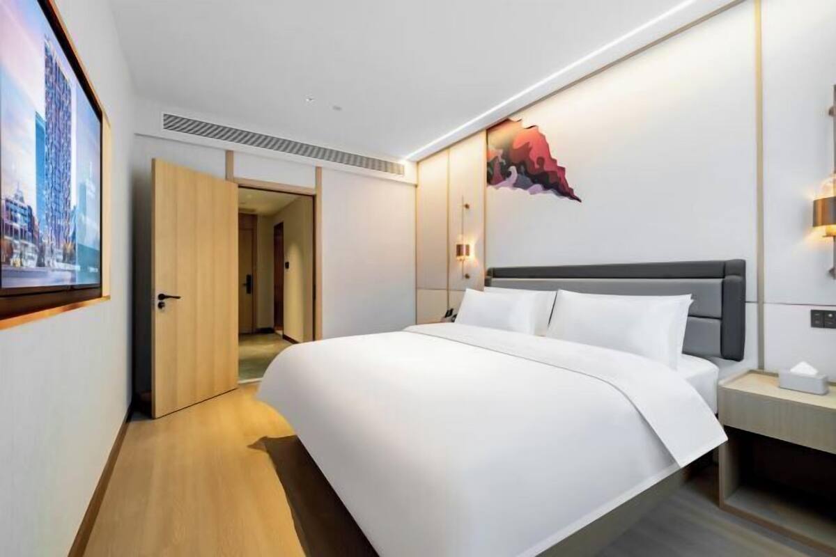 A modern hotel room with a large bed, a wooden door, a painting, and a nightstand.