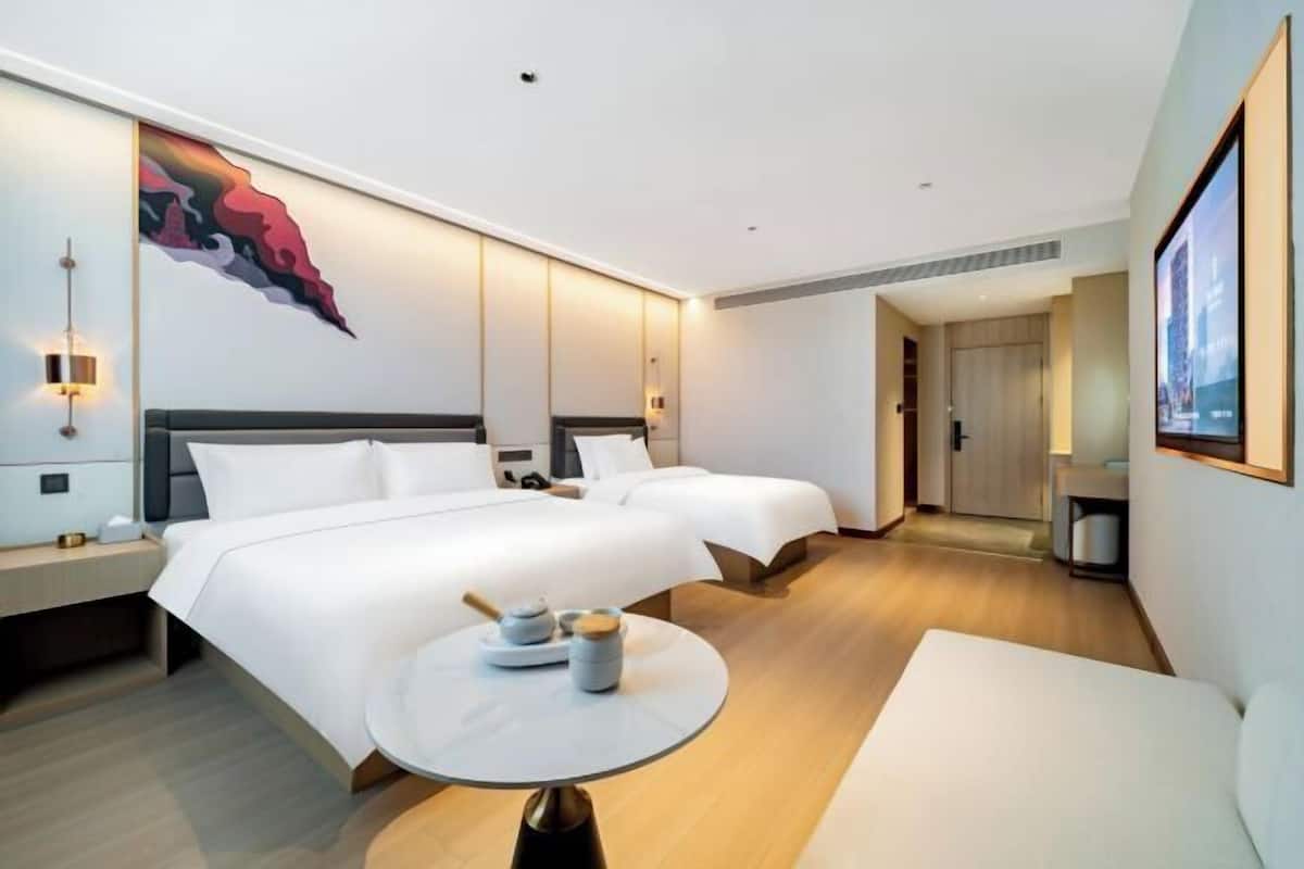 A hotel room with two beds, a small round table, a wall-mounted artwork, and a door leading to another room.