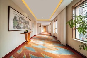 A hotel corridor with a patterned carpet, a framed artwork on the wall, a potted plant, and a window with curtains. - Elong Wanguo Hotel (Liaoyuan Railway Station Traditional Chinese Medicine Hospital) (Liaoyuan)