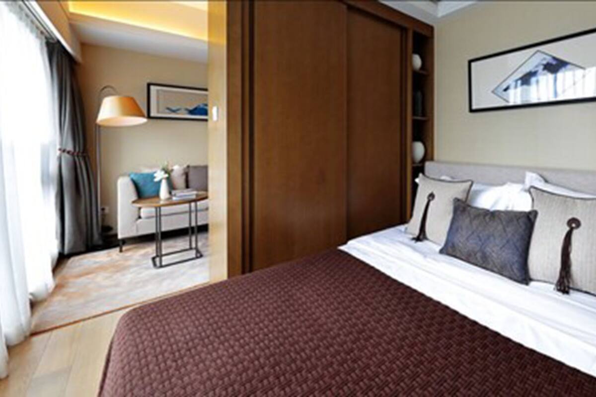A hotel room with a large bed, a desk, and a chair. There is a window with curtains, a framed picture on the wall, and a wooden wardrobe.